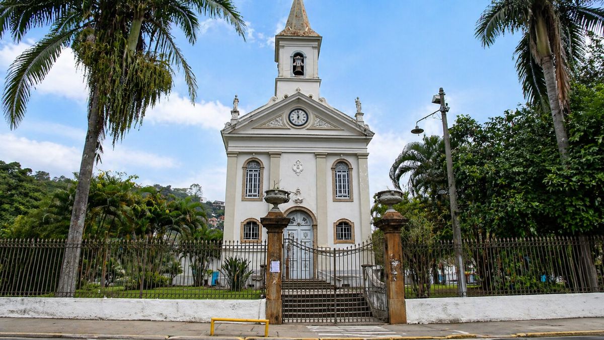 Fotografia frontal da Igreja Matriz de Santana apresentando uma fachada branca em estilo neoclássico com uma torre sineira centralizada no topo. A estrutura possui um frontão triangular com um relógio circular e é emoldurada por palmeiras imperiais altas em ambos os lados. Em primeiro plano observa-se um portão de ferro preto entre pilares de pedra com vasos ornamentais e um jardim gramado que separa a edificação da calçada sob um céu claro com nuvens esparsas.