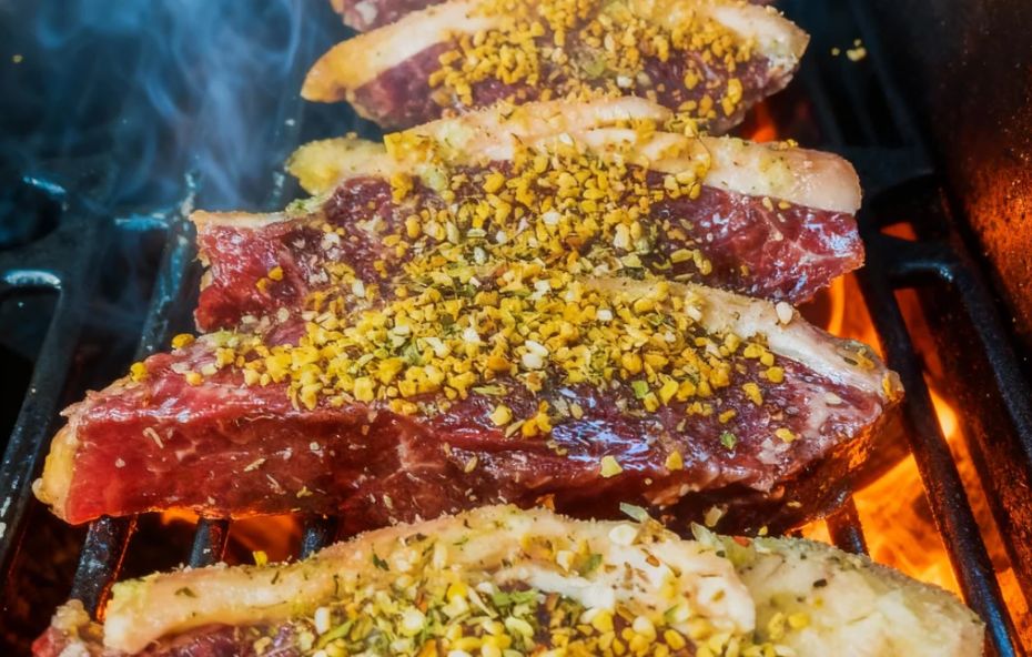 Quatro fatias espessas de picanha crua com capa de gordura branca, cobertas com tempero granulado amarelo e ervas, dispostas sobre uma grelha de metal com chamas altas e brasas vivas ao fundo.