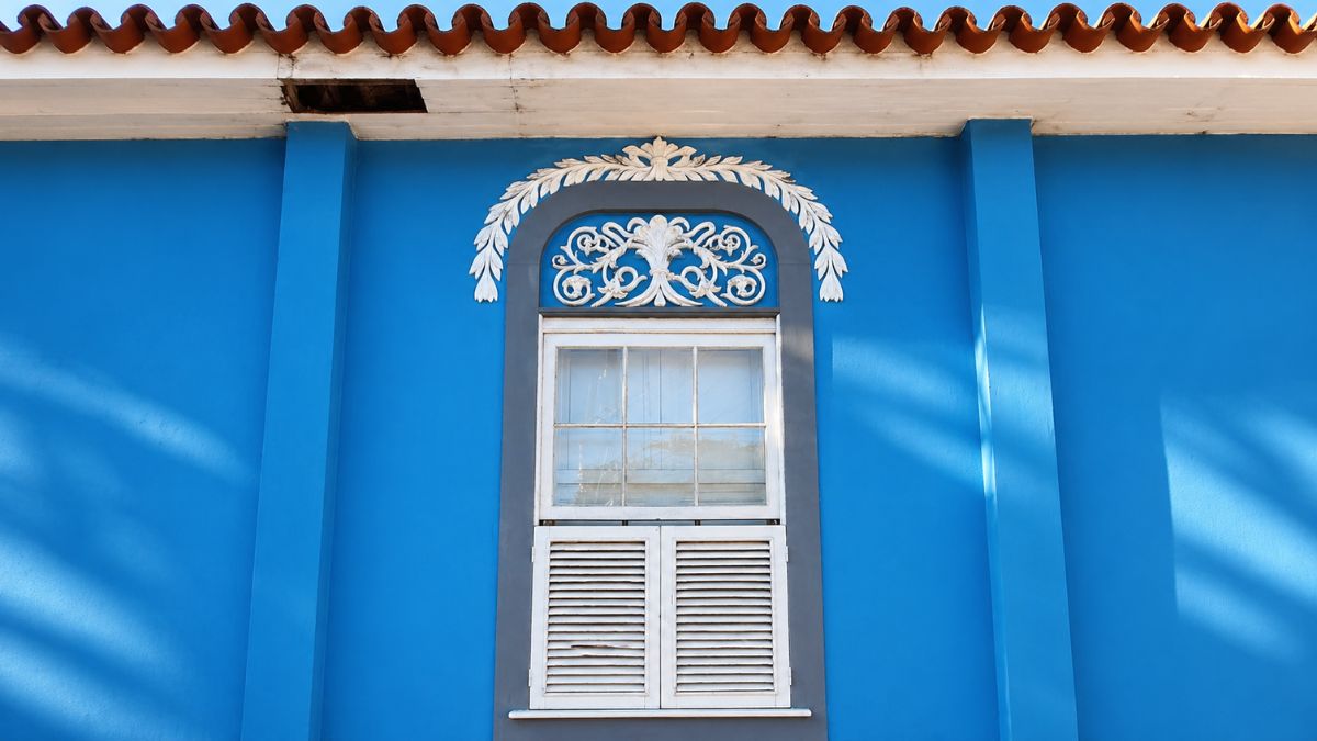 Fotografia em close de uma janela colonial centralizada em uma parede azul vibrante. A parte superior da janela possui um arco decorado com guirlandas brancas esculpidas em alto relevo e um ornamento central em formato de leque. A janela apresenta uma moldura cinza, vidraças quadradas na parte de cima e venezianas brancas fechadas na parte de baixo, posicionada logo abaixo de um beiral de telhas cerâmicas avermelhadas.