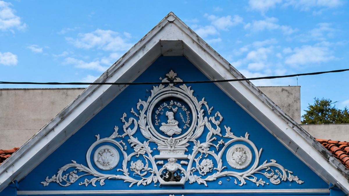 Fotografia em close de um frontão triangular azul vibrante decorado com ricos ornamentos em alto relevo branco. No centro destaca-se um medalhão circular com a efígie da Princesa Isabel cercado por ramos folheados e volutas simétricas. Abaixo do medalhão central existem dois círculos menores com figuras heráldicas interligados por guirlandas de flores e ramos que se estendem pelas laterais sobre o fundo azul e sob um céu com nuvens leves.