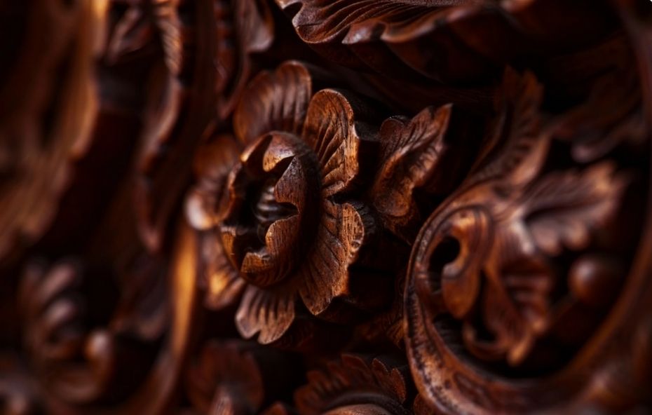 Close de um entalhe em madeira escura com motivos florais em alto relevo demonstrando a precisão técnica e os detalhes das pétalas e folhas esculpidas à mão por artesãos locais.