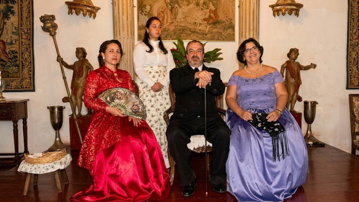 Quatro pessoas posam com trajes de época em um salão clássico, incluindo vestidos luxuosos em vermelho e lilás e terno.