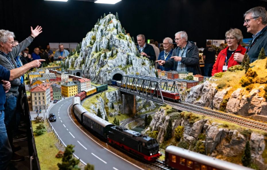 Pessoas de diversas idades observando e apontando para uma grande maquete ferroviária detalhada que contém montanhas com neve, túneis, pontes metálicas e trens em movimento por uma vila em miniatura.