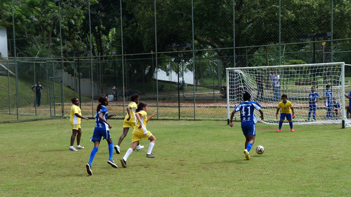 Meninas adolescentes jogando futebol em um campo gramado com trave branca e alambrado ao fundo durante atividade esportiva institucional.