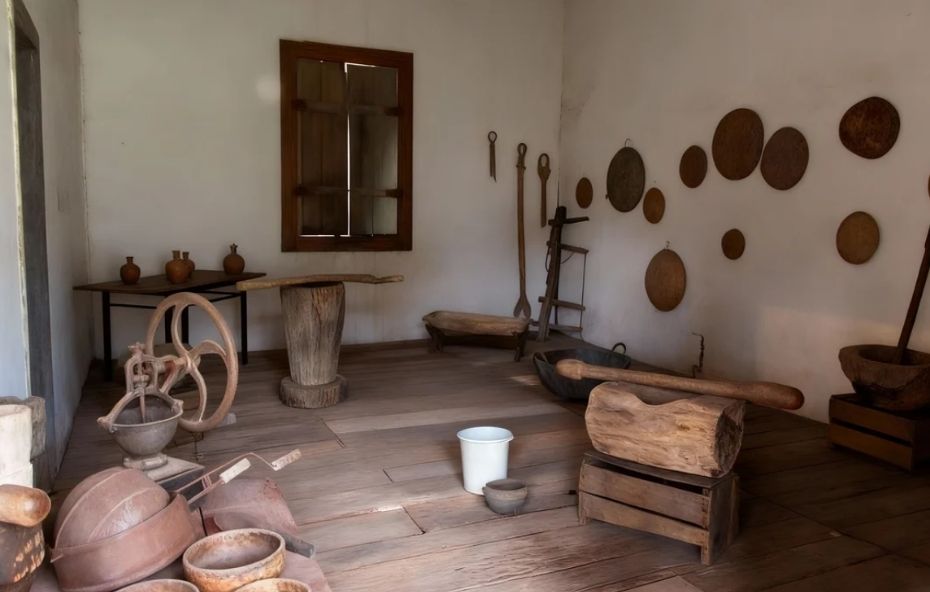 Fotografia do interior de uma sala de museu com piso de madeira clara contendo diversos objetos históricos, como pilões de madeira, rodas de moagem, vasos de barro sobre uma mesa e peneiras circulares penduradas em uma parede branca ao fundo.