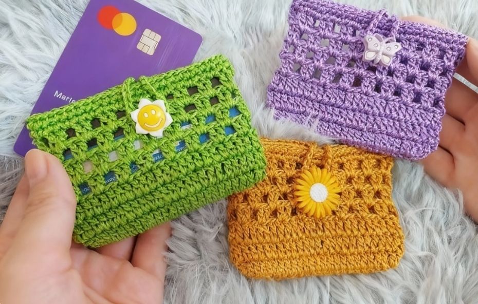 Mãos segurando três pequenos porta cartões feitos de crochê nas cores verde, roxo e mostarda com detalhes de flores e borboleta plástica sobre fundo de pelúcia cinza.