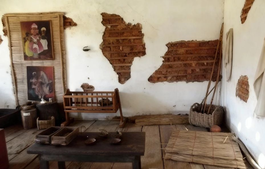 Fotografia do interior de uma sala com paredes de pau a pique revelando a estrutura de ripas de madeira e barro, contendo um berço antigo, quadros com temática afro-brasileira, cestos de palha e uma mesa de madeira escura com utensílios.