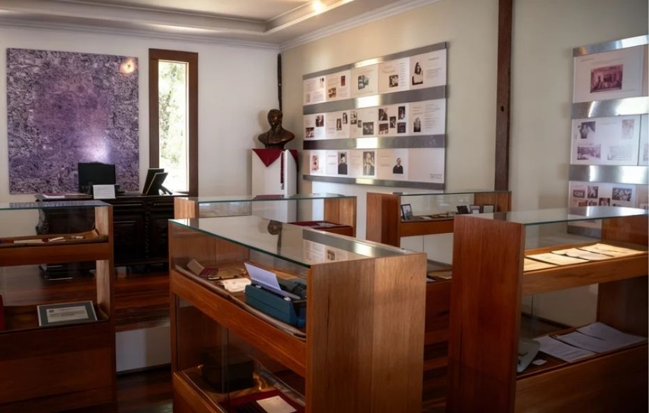 Fotografia do interior de uma sala de exposição com vitrines de madeira e vidro contendo documentos e objetos históricos, além de painéis informativos na parede e um busto de bronze ao fundo.