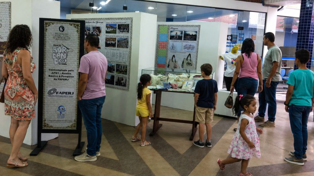 Visitantes adultos e crianças observam painéis informativos e vitrines com bolsas bordadas do projeto Bordando o Vale em um saguão amplo.