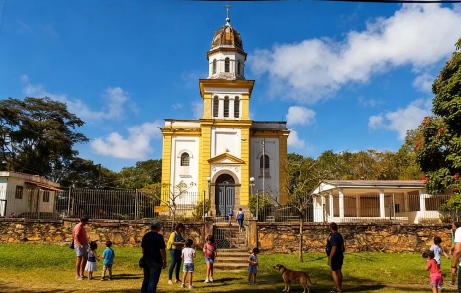 Fotografia da fachada frontal da Igreja Nossa Senhora da Piedade em Ipiabas com pintura em branco e amarelo exibindo uma torre central escalonada com cúpula e cruz no topo em um dia ensolarado com pessoas no gramado frontal.