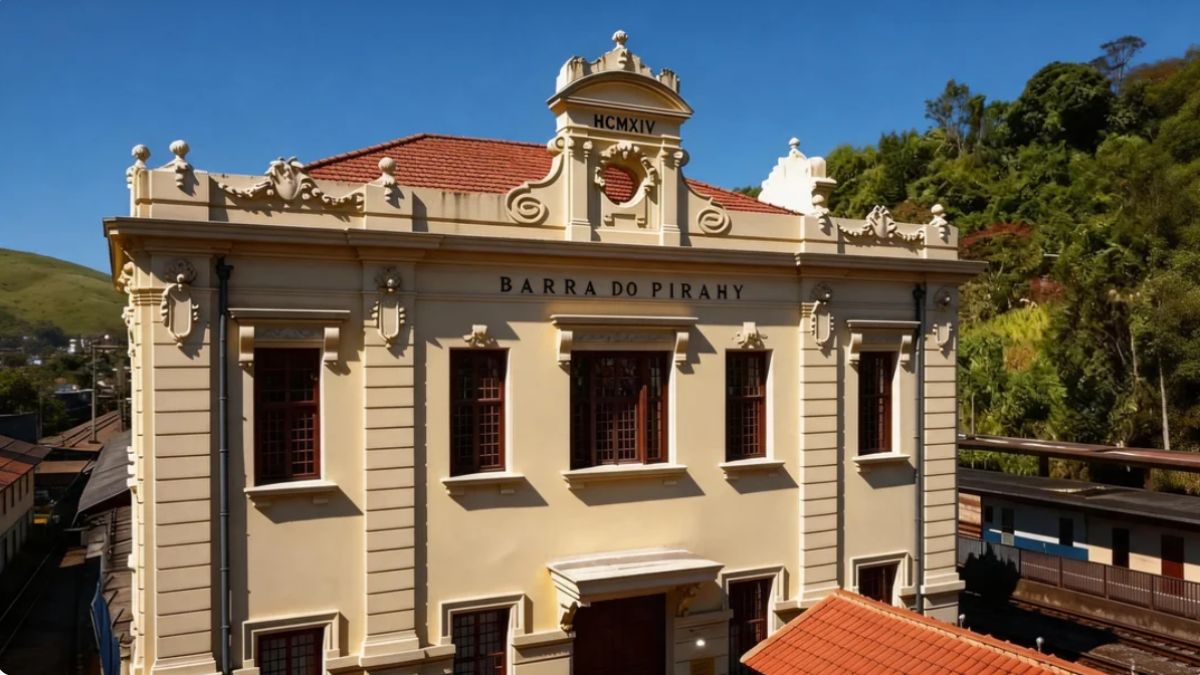 Fotografia da fachada frontal do Centro Cultural Rosemar Muniz Pimentel em Barra do Piraí. O prédio possui arquitetura neoclássica em tom bege com detalhes ornamentados no topo, exibindo a inscrição Barra do Pirahy. Ao fundo, observa-se vegetação verde e trilhos ferroviários sob um céu azul limpo.