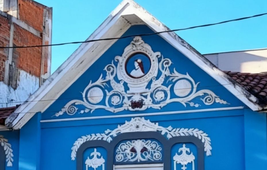Fotografia de uma edificação histórica de esquina pintada em azul claro com detalhes arquitetônicos em branco e telhado de barro marrom. A estrutura apresenta janelas de madeira com venezianas cinzas e molduras ornamentadas por guirlandas em alto relevo. No frontão triangular elevado destaca-se um medalhão central com o busto da Princesa Isabel cercado por arabescos brancos sobre fundo azul.