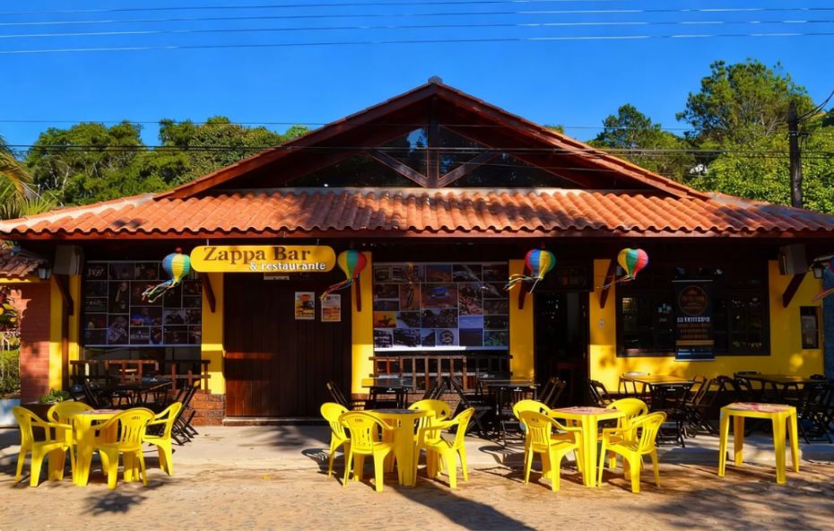 Vista frontal de um restaurante de estilo rústico com paredes amarelas, telhado colonial de barro e mesas externas com cadeiras amarelas sob a luz do sol.