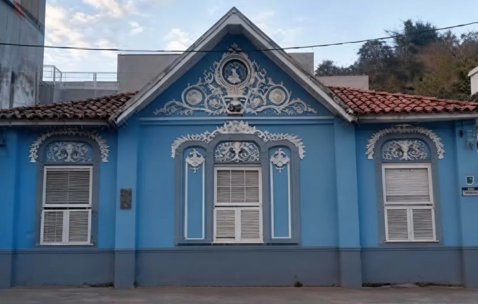 Fotografia da fachada de uma casa histórica pintada em azul claro com rodapé cinza e detalhes arquitetônicos brancos. A imagem mostra janelas de madeira com venezianas brancas e molduras superiores decoradas com guirlandas em alto relevo. No frontão triangular centralizado sob o telhado de telhas cerâmicas escuras destaca-se um medalhão circular com o busto da Princesa Isabel em perfil cercado por ornamentos de arabescos brancos.