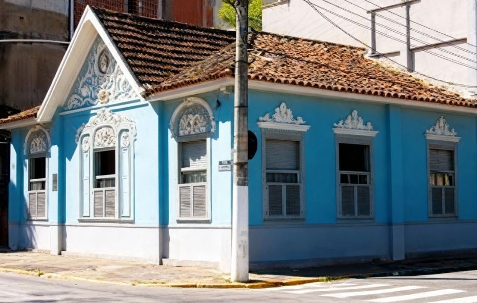 Fotografia de uma casa histórica de esquina pintada em azul vibrante com detalhes ornamentais em branco. O prédio possui um frontão triangular elevado com guirlandas e molduras decorativas acima das janelas cinzas. A calçada contorna o imóvel em uma área urbana com faixa de pedestres no asfalto e um poste de concreto posicionado à esquerda da construção.