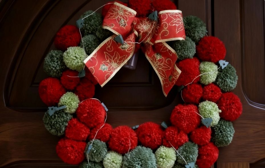 Guirlanda decorativa artesanal feita com pompons de lã nas cores vermelha, verde escuro e verde claro, adornada com um laço vermelho de estampa dourada e pequenas luzes de LED sobre uma porta de madeira escura.