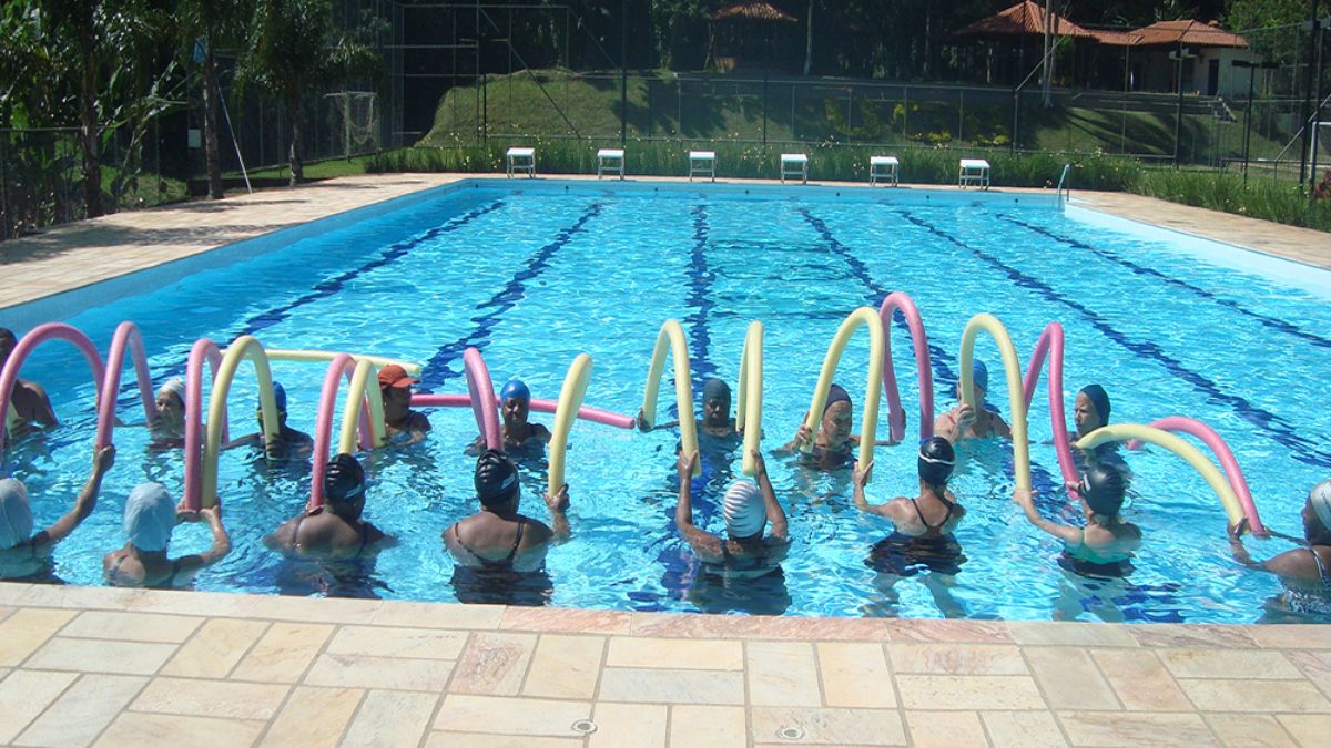 Um grupo de idosos realiza exercícios de hidroginástica com espaguetes coloridos em uma piscina ao ar livre cercada por vegetação.