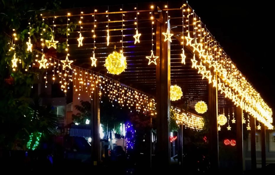 Uma estrutura de pérgola de madeira à noite totalmente decorada com diversas fileiras verticais de luzes amarelas cintilantes e enfeites em formato de estrelas e globos luminosos suspensos sob um céu escuro.
