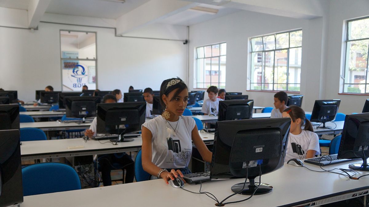 Uma jovem estudante utilizando um computador em uma sala de informática repleta de alunos vestindo a camiseta do projeto institucional.