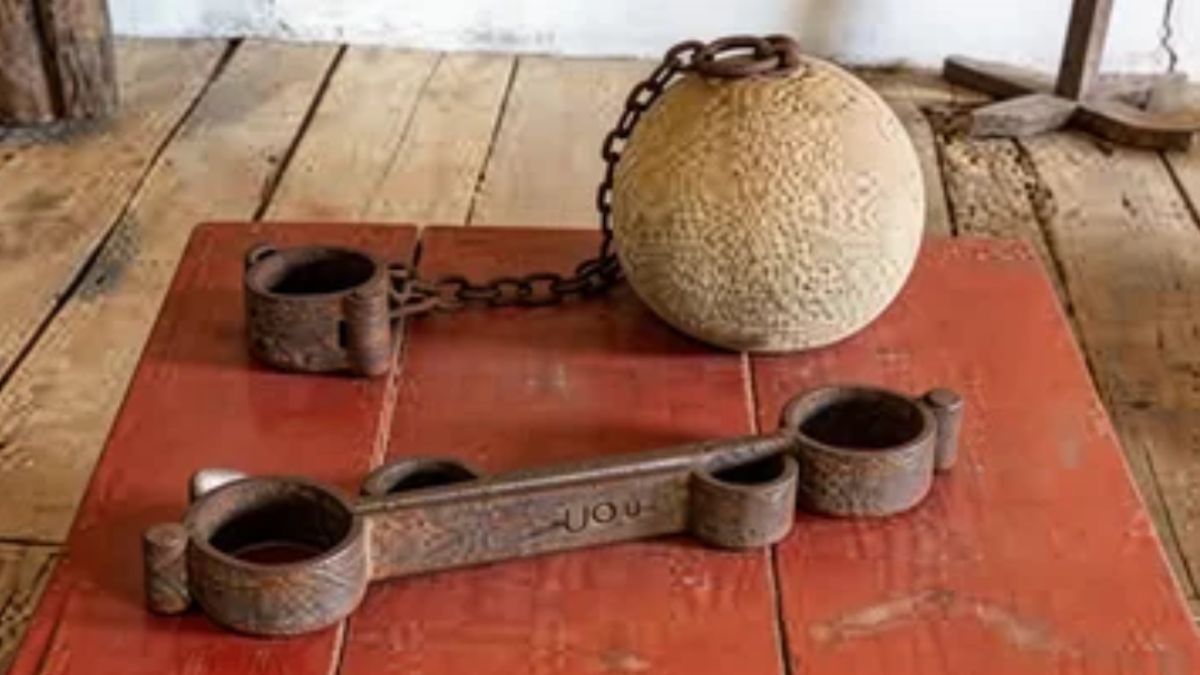 Algemas de ferro e uma grande bola de pedra presas por correntes sobre uma mesa vermelha em ambiente histórico.