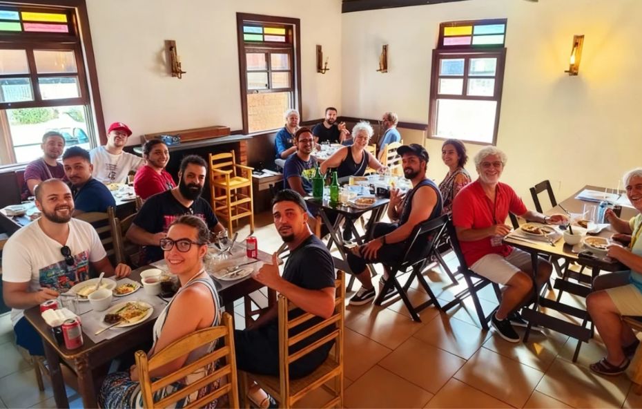 Pessoas sorridentes sentadas em mesas de madeira dentro de um restaurante com paredes brancas e janelas decoradas com vidros coloridos. O ambiente é iluminado por arandelas clássicas nas paredes e possui um piso cerâmico em tom ocre.