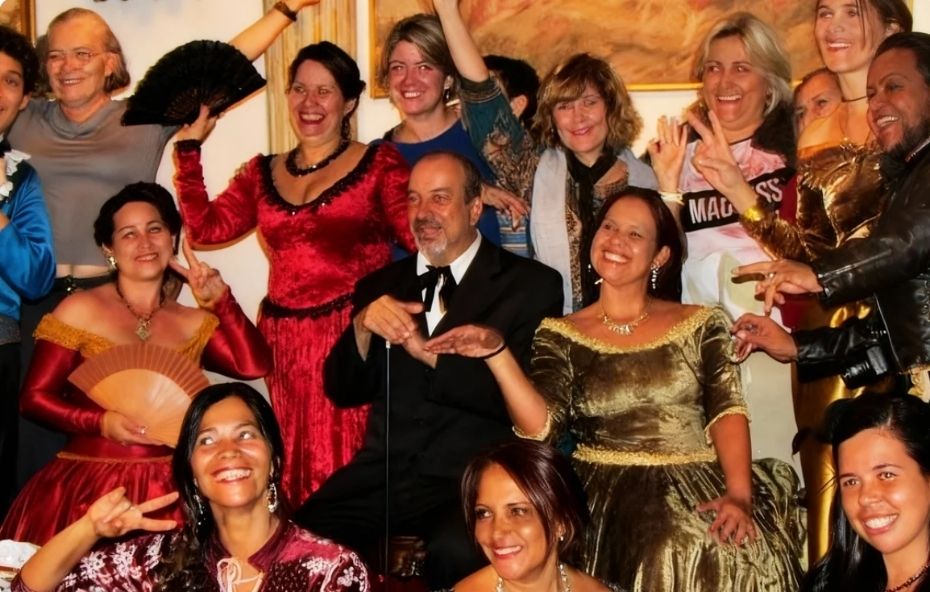 Fotografia em grupo com várias pessoas sorridentes vestindo roupas clássicas do século dezenove como vestidos longos de veludo com decotes rendados e um homem ao centro com terno preto e gravata borboleta em um ambiente interno iluminado.