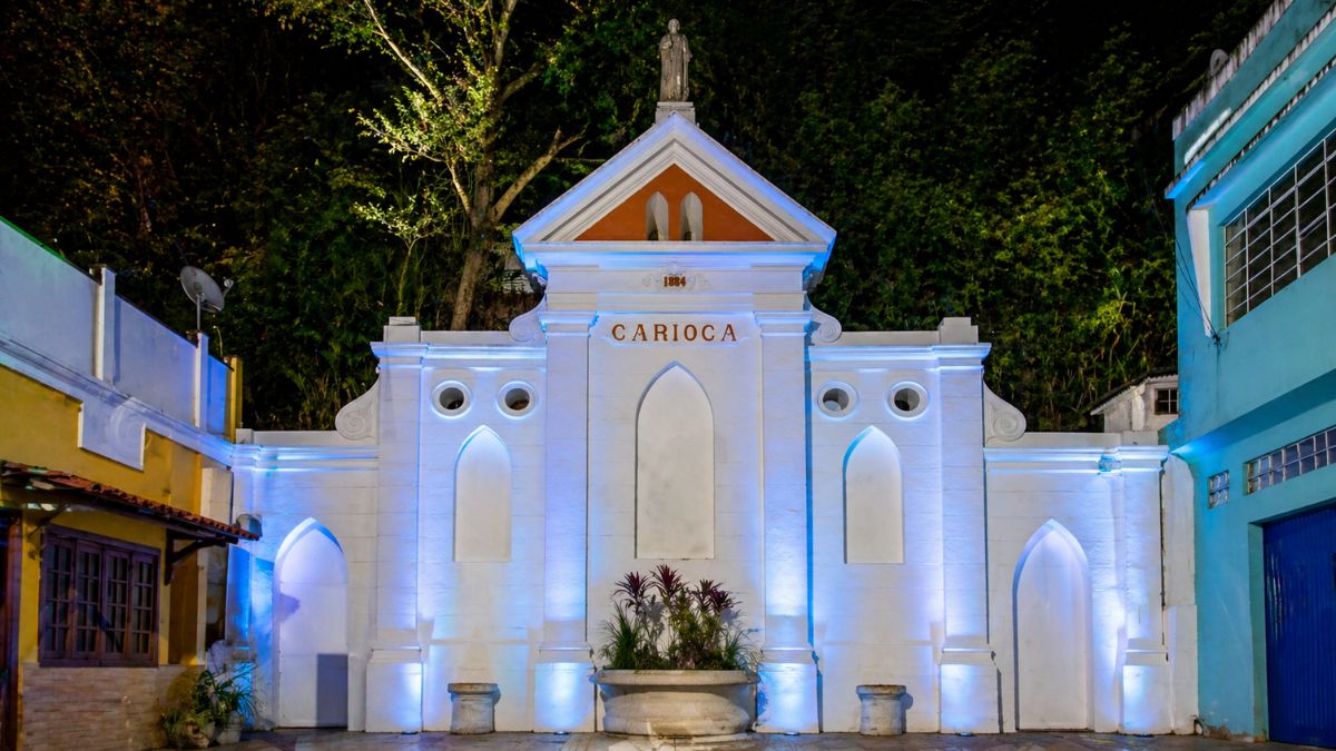 Fotografia noturna do Chafariz da Carioca com iluminação artística em tons de azul e branco. A estrutura de alvenaria branca apresenta um estilo neoclássico com nichos em arco ogival e um frontão triangular central que ostenta a inscrição Carioca e o ano mil oitocentos e oitenta e quatro. Uma estátua pequena repousa no topo do monumento enquanto a vegetação densa e escura serve de fundo para a construção iluminada que possui um vaso central com folhagens na base.