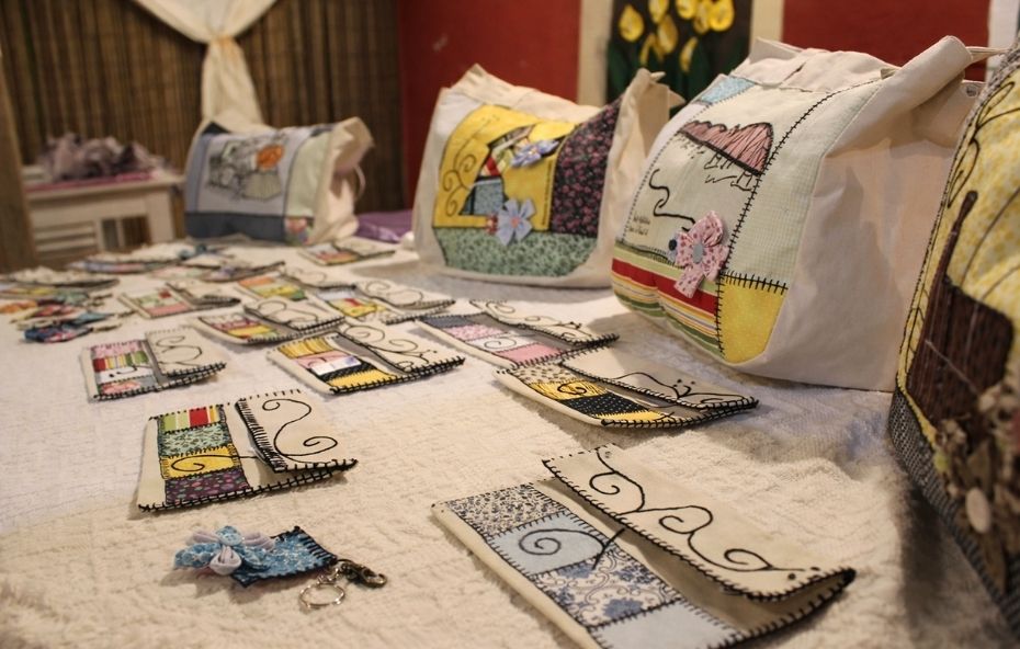 Fotografia em close de diversas bolsas e carteiras de tecido artesanais dispostas sobre uma mesa branca, apresentando bordados detalhados de flores e elementos arquitetônicos coloridos em estilo rústico.