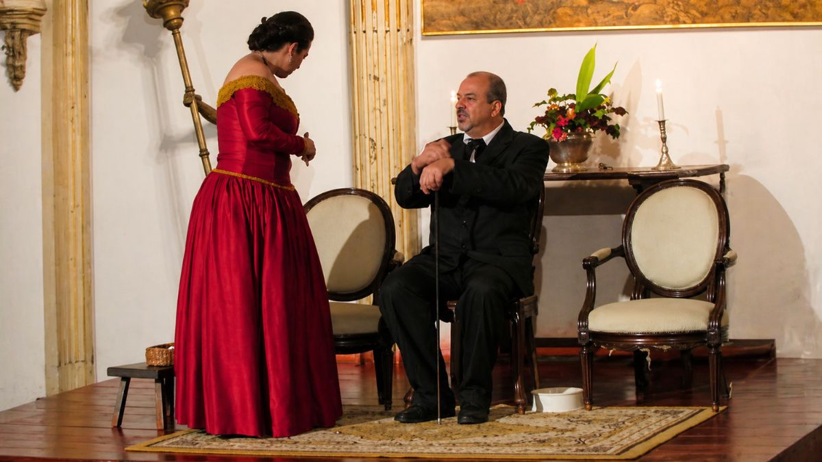 Um homem de terno preto sentado e uma mulher com vestido longo carmesim interagem em um cenário de época iluminado.