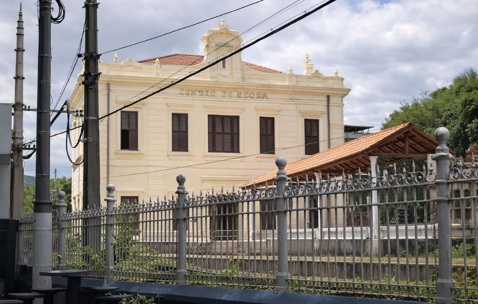 Fotografia capturada por trás de uma grade metálica cinza, mostrando o edifício histórico de cor clara e uma plataforma de embarque com telhado de barro sob um céu nublado.