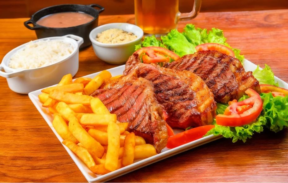 Prato branco com fatias de picanha grelhada sobre folhas de alface e tomate, acompanhado de uma porção generosa de batatas fritas douradas e tigelas com arroz, farofa e feijão.