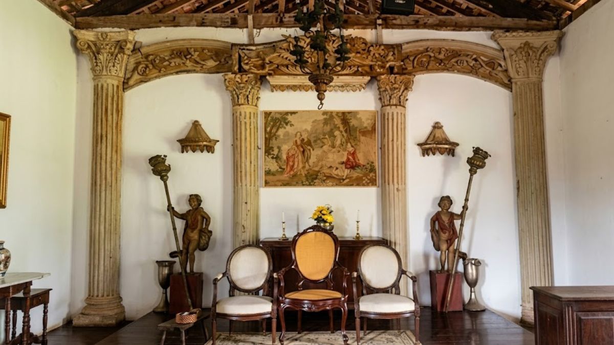 Cenário clássico com colunas esculpidas, teto de madeira aparente, cadeiras de época sobre tapete e estátuas ornamentais em ambiente preservado.