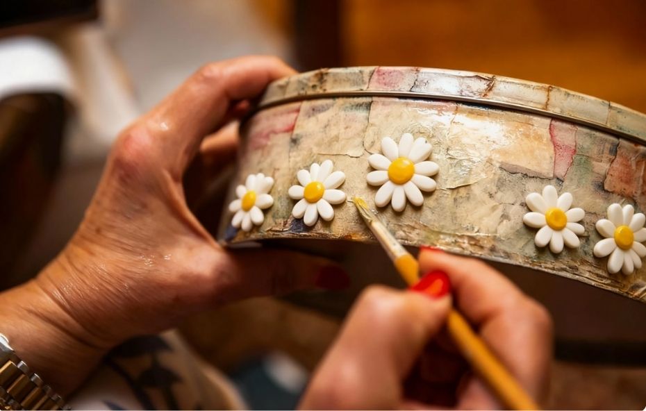 Mãos de uma artesã com unhas vermelhas segurando um pincel fino para aplicar detalhes em uma caixa redonda decorada com técnica de decoupage e aplicações de flores brancas.