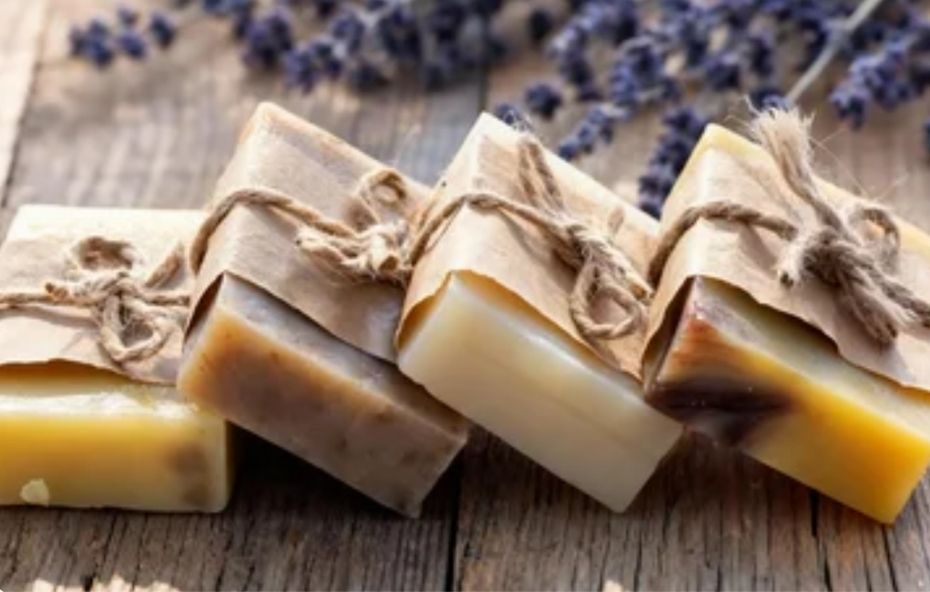 Quatro barras de sabonete artesanal com texturas naturais dispostas em uma tábua de madeira rústica ao lado de um ramo de lavanda roxa fresca.