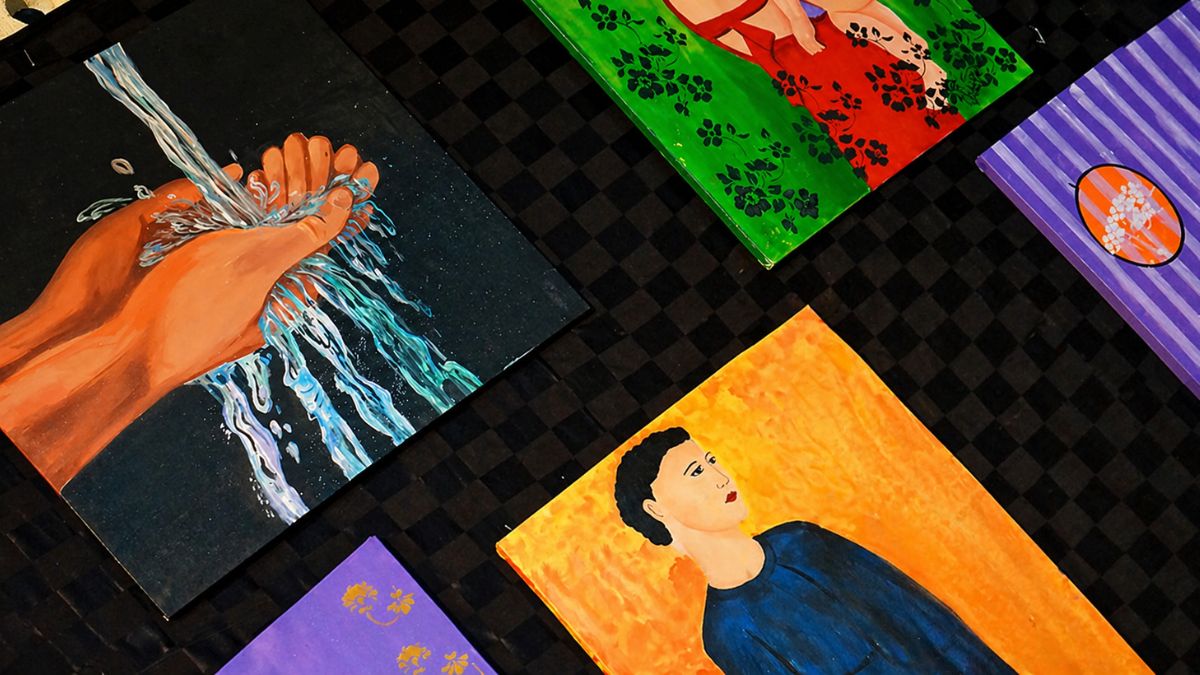 Vista superior de cinco telas de pintura coloridas dispostas sobre uma superfície quadriculada escura na Galeria de Exposições da instituição.