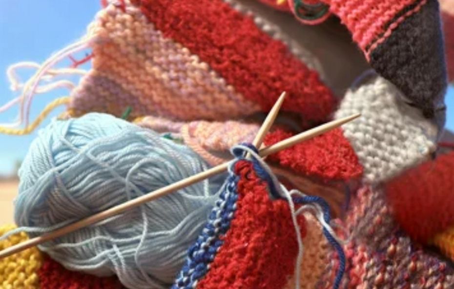 Close em agulhas de tricô de madeira cruzadas iniciando um trabalho com linha azul e vermelha sobre um fundo de quadrados de lã coloridos em exposição ao ar livre.