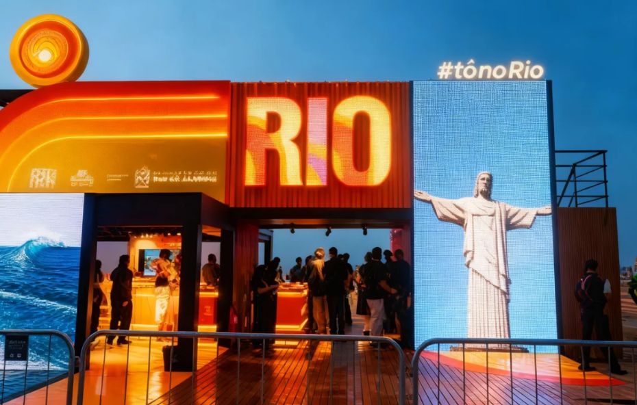 Estrutura moderna composta por contêineres coloridos com painéis de LED exibindo a imagem do Cristo Redentor e a hashtag Tô no Rio enquanto pessoas visitam o estande ao entardecer.