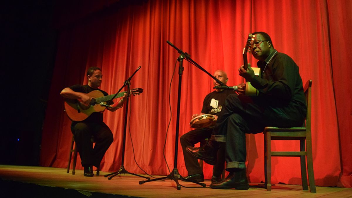 Três músicos homens apresentam-se em um palco com violões e pandeiro diante de uma grande cortina vermelha no Centro Cultural Aracy Carvalho Di Biase.