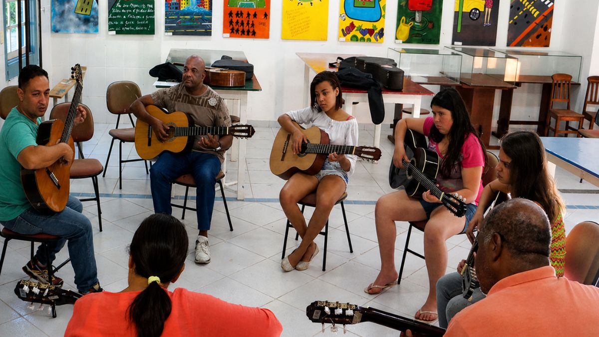 Um grupo diversificado de pessoas sentadas em círculo pratica violão em uma sala de artes decorada com quadros coloridos no Centro Cultural.