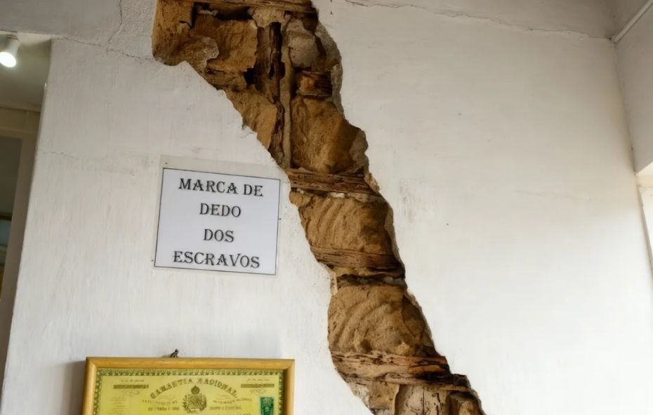 Fotografia de uma parede branca com uma grande fissura vertical proposital, que revela a estrutura interna de barro, ripas de madeira e pedras, exibindo ao lado uma placa com a inscrição marca de dedo dos escravos.