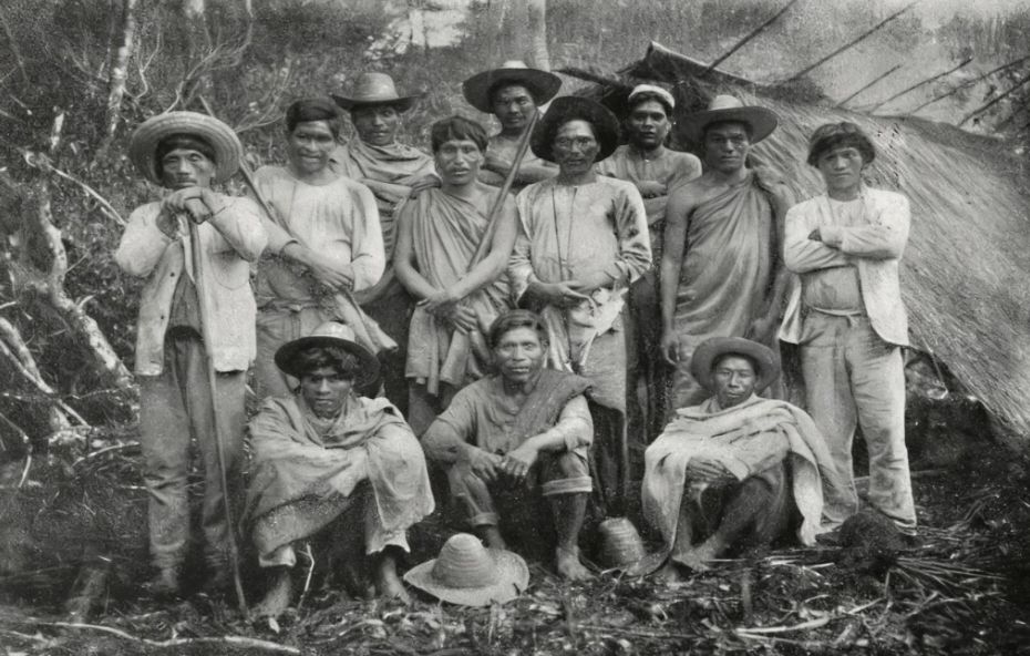 Gravura colorida do século dezenove mostrando uma aldeia indígena com malocas circulares em meio à densa vegetação da Mata Atlântica e nativos com corte de cabelo em formato de coroa.