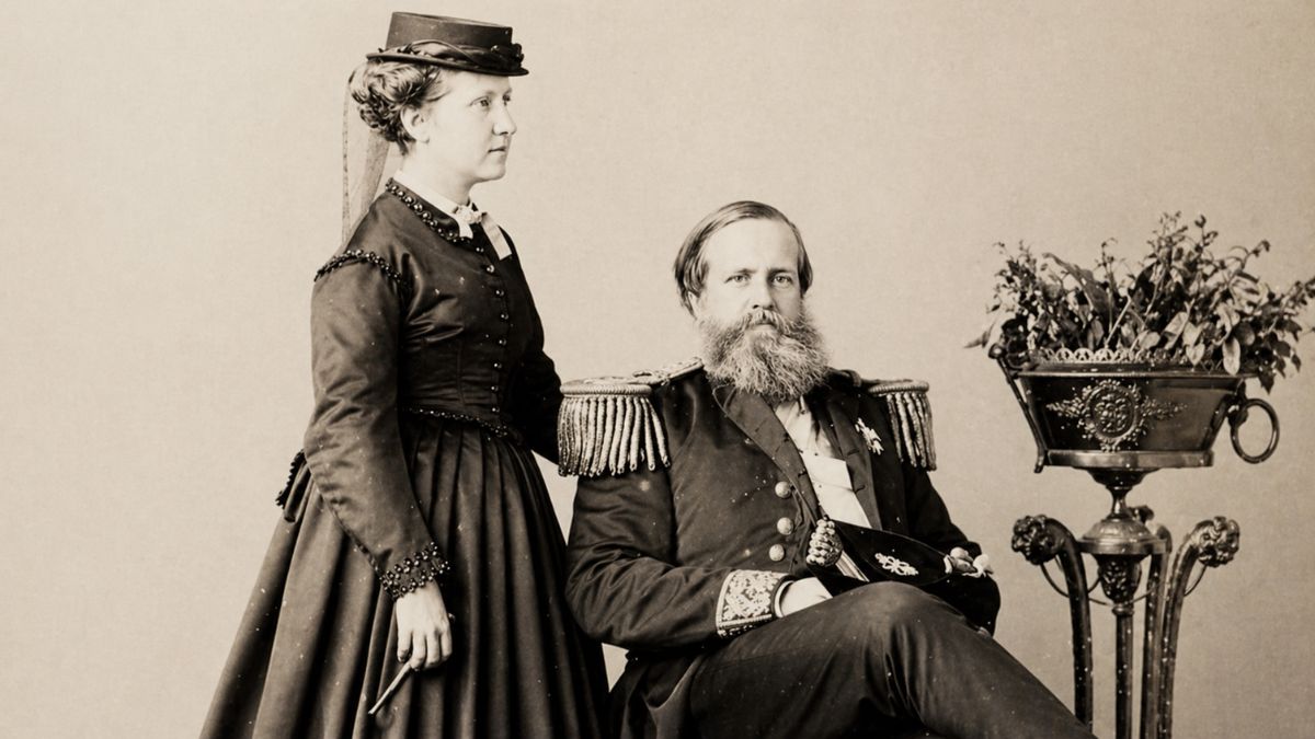 Fotografia antiga em tons de sépia apresentando a Princesa Isabel e o Imperador Dom Pedro Segundo. À esquerda a princesa aparece de pé e de perfil vestindo um traje escuro com detalhes em pedrarias e um chapéu pequeno. Ao centro o imperador está sentado de frente usando sua farda militar com dragonas nos ombros e ostentando uma barba longa e clara. Ao lado direito observa-se um vaso ornamental com plantas sobre um suporte metálico trabalhado.