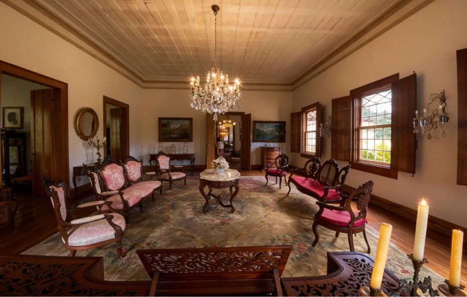 Fotografia do interior de uma sala histórica na Fazenda Cachoeira Grande exibindo sofás em tom rosa antigo com estampas florais e cadeiras clássicas distribuídas sobre um tapete ornamentado sob um grande lustre de cristal pendente do teto de madeira.