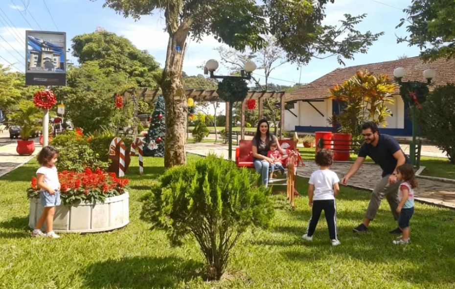 Família com três crianças pequenas brincando em um gramado ensolarado na praça central de Ipiabas. Ao fundo há decorações natalinas, árvores e uma construção histórica colonial branca com detalhes azuis sob um céu claro.