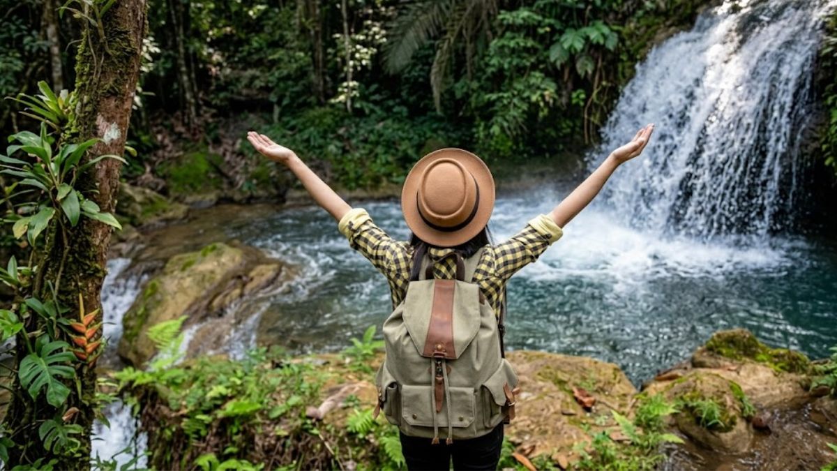 Vista traseira de uma mulher com chapéu e mochila bege de braços abertos diante de uma cachoeira em Ipiabas.