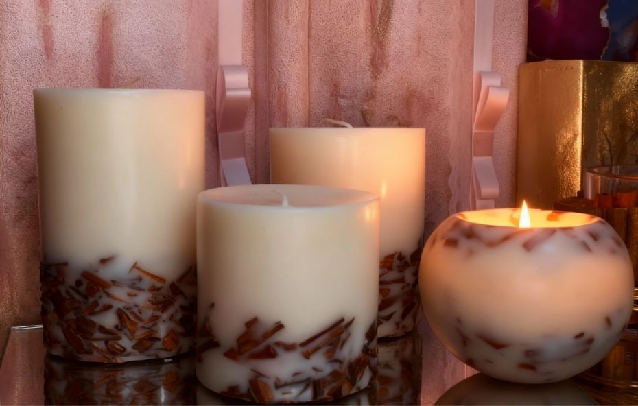 Conjunto de quatro velas artesanais na cor creme com fragmentos de madeira natural incrustados na base. Uma das velas é redonda e está acesa sobre uma superfície espelhada.