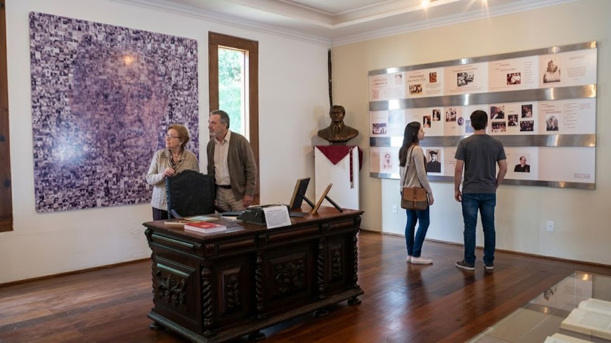 Visitantes exploram a sala de exposição do Centro Cultural Aracy Carvalho Di Biase, observando o escritório histórico e os painéis biográficos.