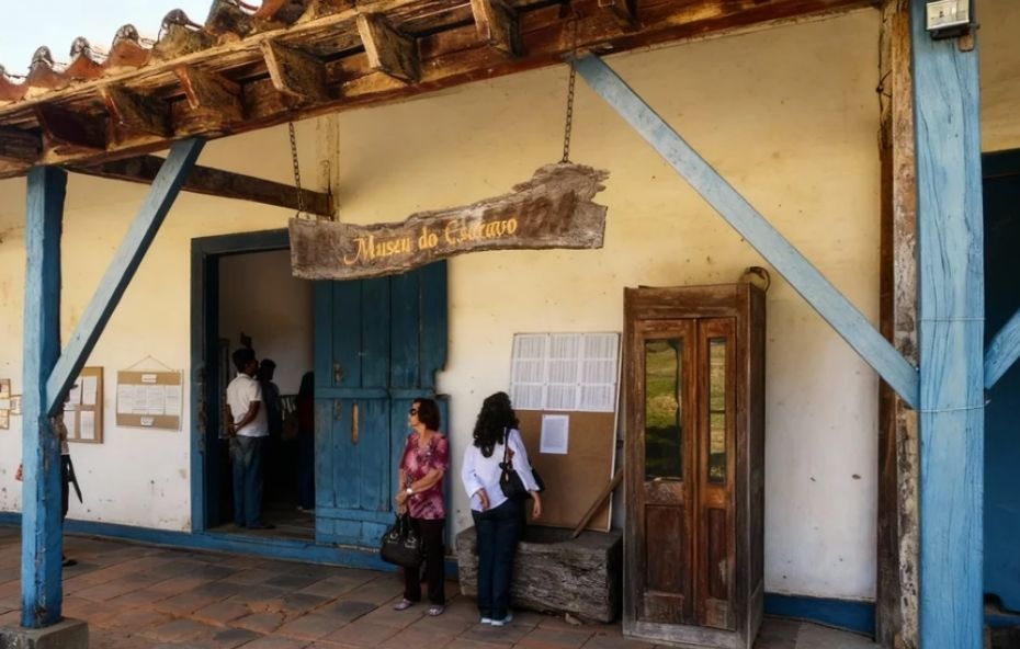 Fotografia da fachada de uma construção histórica com telhado de barro e vigas de madeira onde duas mulheres observam um painel informativo ao lado de uma porta azul larga e uma placa de madeira suspensa com o nome do museu.