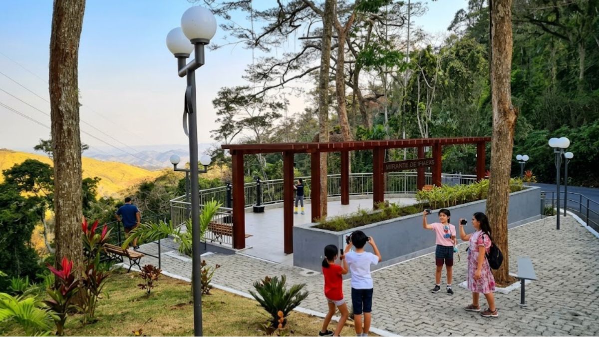 Fotografia capturando um mirante ao ar livre com um grupo de pessoas, incluindo crianças, tirando fotos no primeiro plano, e uma vista panorâmica de colinas e florestas ao fundo, com uma estrutura de madeira e luminárias de poste.