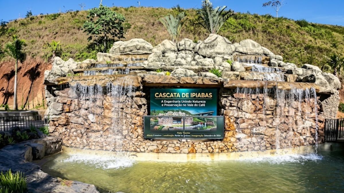 Fotografia colorida da Cascata de Ipiabas mostrando a queda d’água sobre rochas artificiais com placa informativa central sob céu azul.